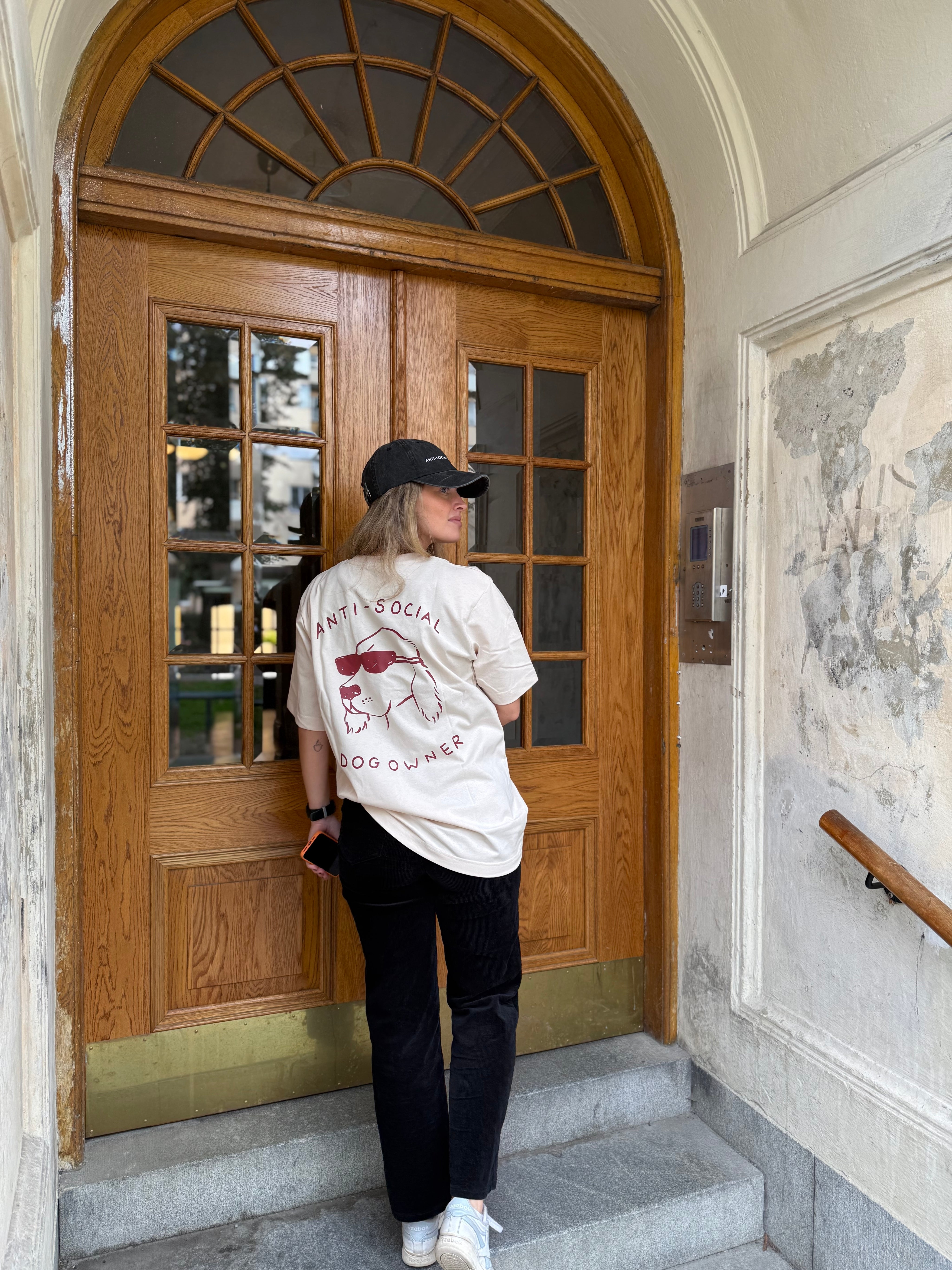 Person wearing a white t-shirt with red text and graphics, black pants, and a black cap standing in front of a wooden door.